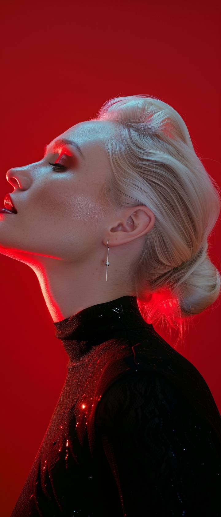 A side profile shot of a model with platinum blonde hair, illuminated by soft, white lighting against a vibrant red background. The makeup is minimal, emphasizing the model’s pale complexion and red lips. The overall effect is striking and minimalist, with a focus on the contrast between the red background and the model’s pale skin.