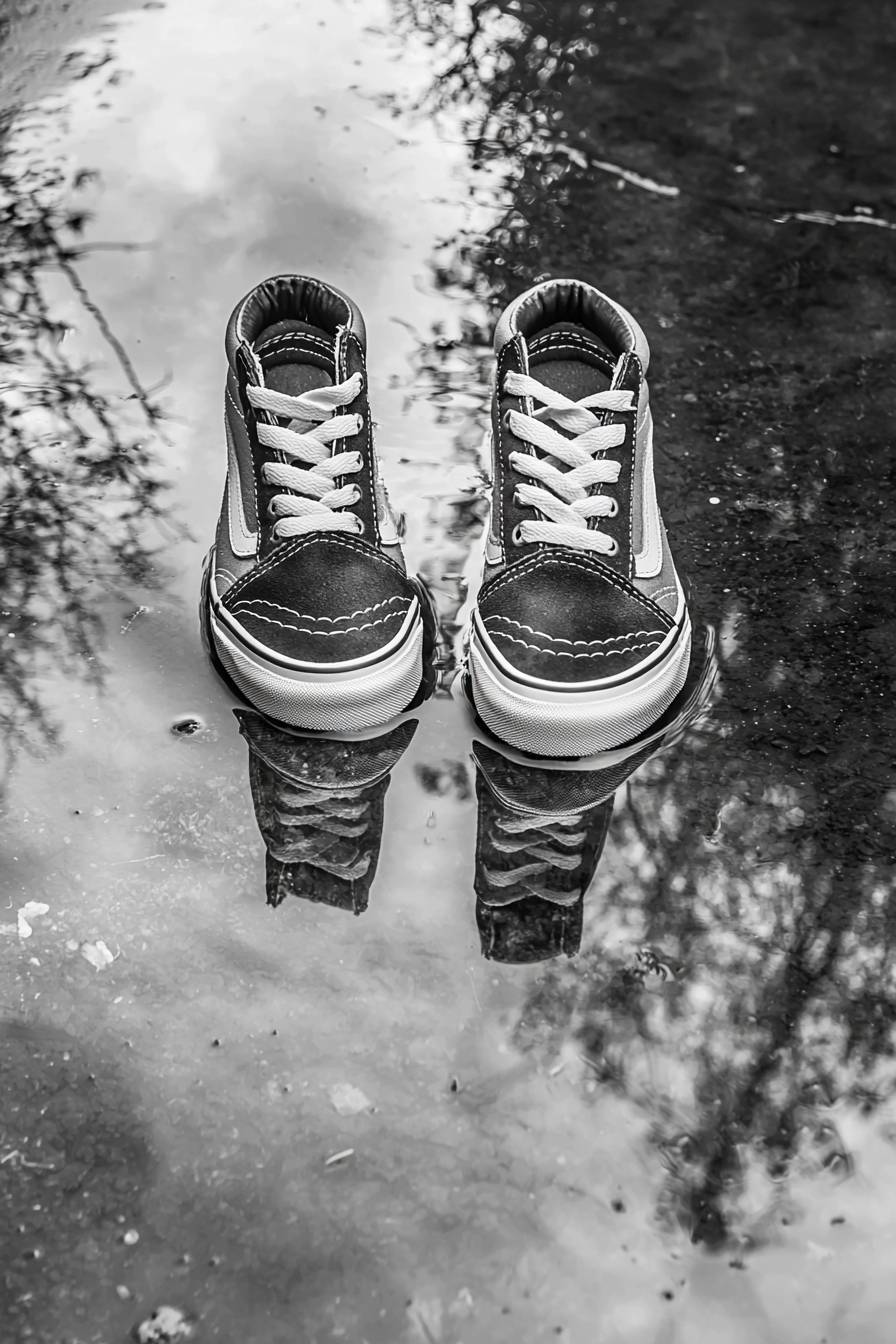 A pair of black and white Vans shoes reflected in a water puddle.