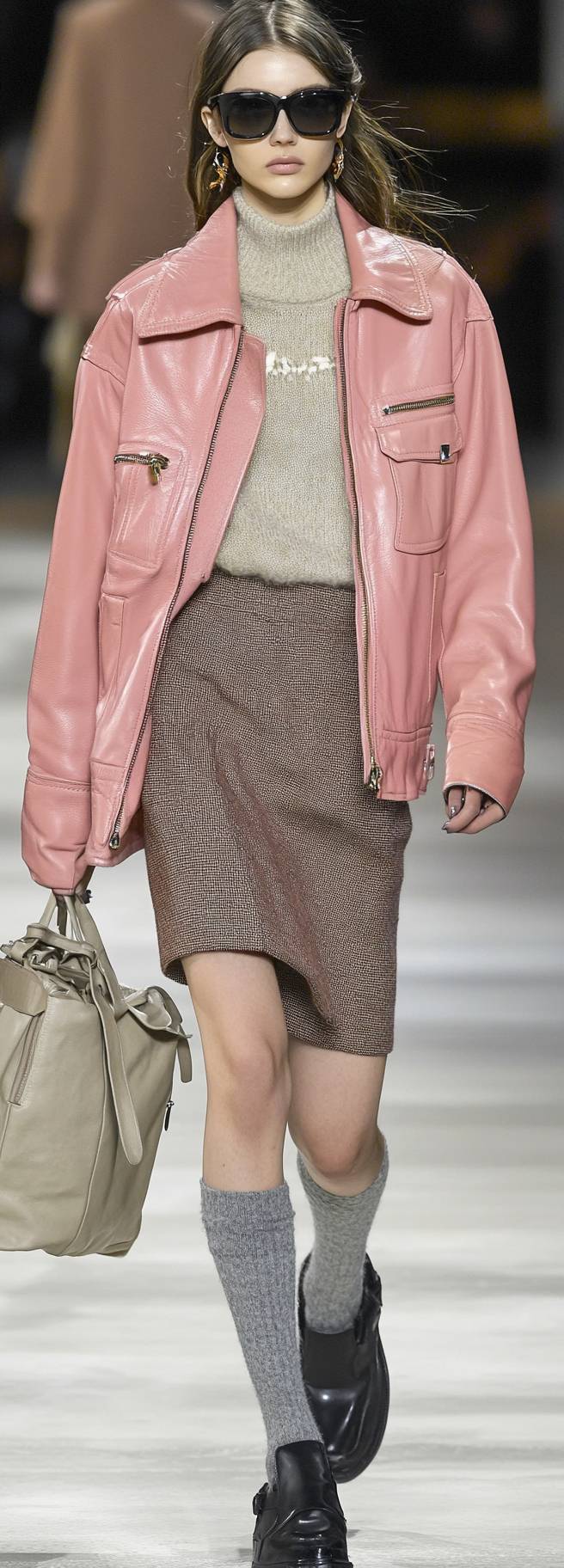 Una modelo camina por la pasarela con una chaqueta de cuero rosa, una falda de lana marrón, calcetines grises y zapatos negros, llevando una bolsa beige y gafas de sol.